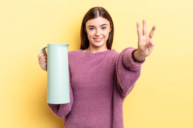 Genç İspanyol kadın gülümsüyor ve arkadaş canlısı görünüyor, üç numarayı gösteriyor. Kahve termosu kavramı