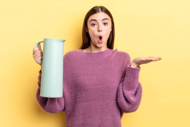 Genç İspanyol kadın şaşırmış ve şok olmuş görünüyor. Çenesi düşmüş ve elinde bir nesne tutuyor. Kahve termosu kavramı