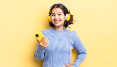 Genç İspanyol kadın mutlu bir şekilde gülümsüyor, eli kalçasında ve kendinden emin. kulaklıklar ve akıllı telefon konsepti