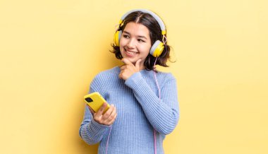 Genç İspanyol kadın mutlu ve kendinden emin bir ifadeyle gülümsüyor. Elleri çenesinde. kulaklıklar ve akıllı telefon konsepti