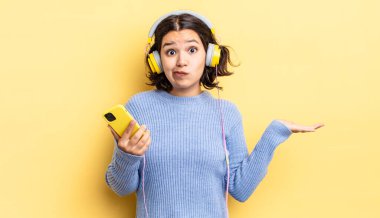 Genç İspanyol kadın şaşkın, şaşkın ve şüpheci hissediyor. kulaklıklar ve akıllı telefon konsepti