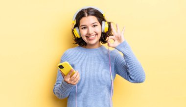 Genç İspanyol kadın kendini mutlu hissediyor, iyi bir jestle onaylıyor. kulaklıklar ve akıllı telefon konsepti