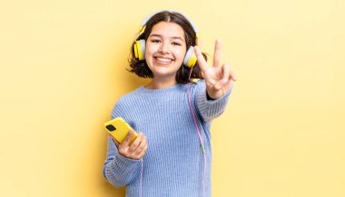 Genç İspanyol kadın gülümsüyor ve mutlu görünüyor, zafer ya da barış işareti yapıyor. kulaklıklar ve akıllı telefon konsepti