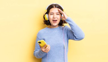 Genç İspanyol kadın mutlu, şaşkın ve şaşırmış görünüyor. kulaklıklar ve akıllı telefon konsepti