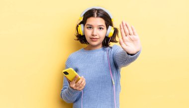 Genç İspanyol kadın ciddi görünüyor. Avuç içini gösteriyor. Durdurma hareketi yapıyor. kulaklıklar ve akıllı telefon konsepti