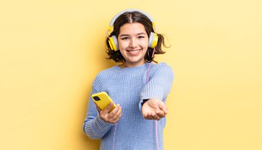 Genç İspanyol kadın mutlu bir şekilde gülümsüyor ve bir konsept sunuyor. kulaklıklar ve akıllı telefon konsepti