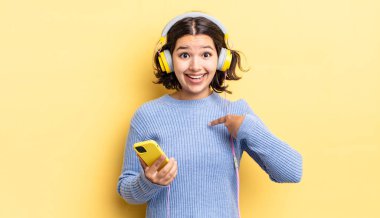 Genç İspanyol kadın kendini mutlu hissediyor ve heyecanla kendini işaret ediyor. kulaklıklar ve akıllı telefon konsepti