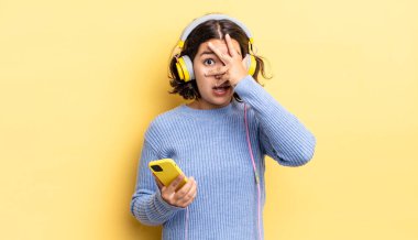 Genç İspanyol bir kadın şok olmuş, korkmuş ya da korkmuş görünüyor. Yüzü elle kapatılmış. kulaklıklar ve akıllı telefon konsepti