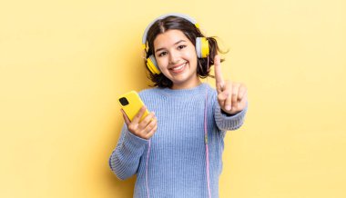 Genç İspanyol kadın gülümsüyor ve arkadaş canlısı görünüyor, bir numarayı gösteriyor. kulaklıklar ve akıllı telefon konsepti