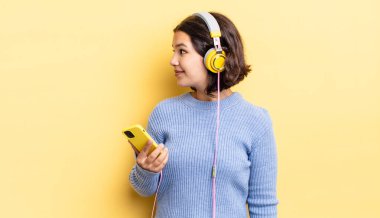 Profil görünümlü genç İspanyol kadın hayal mi kuruyor yoksa hayal mi kuruyor? kulaklıklar ve akıllı telefon konsepti