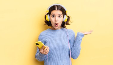 Genç İspanyol kadın şaşırmış ve şok olmuş görünüyor. Çenesi düşmüş ve elinde bir nesne tutuyor. kulaklıklar ve akıllı telefon konsepti