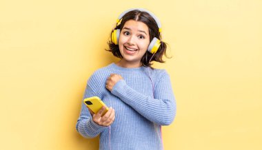 Genç İspanyol kadın kendini mutlu hissediyor ve bir meydan okumayla yüzleşiyor ya da kutluyor. kulaklıklar ve akıllı telefon konsepti