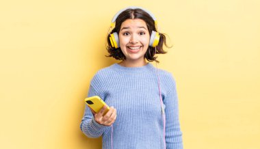 Genç İspanyol kadın mutlu ve hoş bir şekilde şaşırmış görünüyor. kulaklıklar ve akıllı telefon konsepti