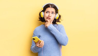 Genç İspanyol kadın düşünüyor, kuşkulu ve şaşkın hissediyor. kulaklıklar ve akıllı telefon konsepti