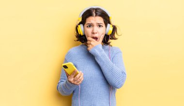 Ağzı ve gözleri sonuna kadar açık ve eli çenesinde olan genç İspanyol kadın. kulaklıklar ve akıllı telefon konsepti