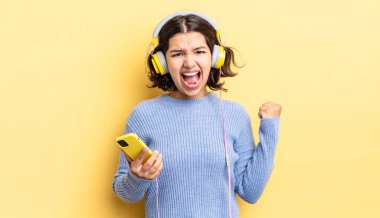 Genç İspanyol kadın öfkeli bir ifadeyle agresif bir şekilde bağırıyor. kulaklıklar ve akıllı telefon konsepti