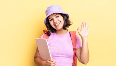 Genç İspanyol kadın mutlu bir şekilde gülümsüyor, el sallıyor, sizi selamlıyor ve selamlıyor. okul konseptine geri dön