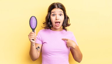 Genç İspanyol kadın şok olmuş ve ağzı açık, kendini işaret ederek şaşırmış görünüyor. saç fırçası konsepti