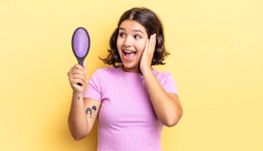 Genç İspanyol kadın mutlu, heyecanlı ve şaşırmış hissediyor. saç fırçası konsepti