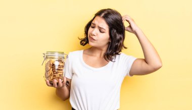 Genç İspanyol kadın şaşkın ve kafası karışık, kaşınan bir kafa hissediyor. kurabiye şişesi konsepti