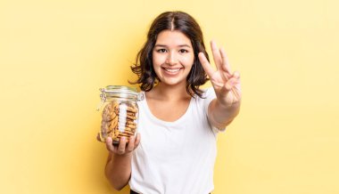 Genç İspanyol kadın gülümsüyor ve arkadaş canlısı görünüyor, üç numarayı gösteriyor. kurabiye şişesi konsepti