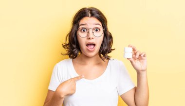 Genç İspanyol kadın şok olmuş ve ağzı açık, kendini işaret ederek şaşırmış görünüyor. hastalık hapları konsepti