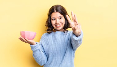 Genç İspanyol kadın gülümsüyor ve arkadaş canlısı görünüyor, üç numarayı gösteriyor. boş kase kavramı