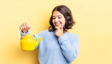 Genç İspanyol kadın mutlu ve kendinden emin bir ifadeyle gülümsüyor. Elleri çenesinde. çaydanlık konsepti