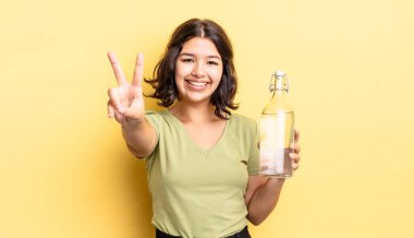 Genç İspanyol kadın gülümsüyor ve arkadaş canlısı görünüyor, iki numarayı gösteriyor. su şişesi kavramı