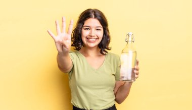 Genç İspanyol kadın gülümsüyor ve arkadaş canlısı görünüyor, dört numarayı gösteriyor. su şişesi kavramı