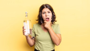 Ağzı ve gözleri sonuna kadar açık ve eli çenesinde olan genç İspanyol kadın. su şişesi kavramı