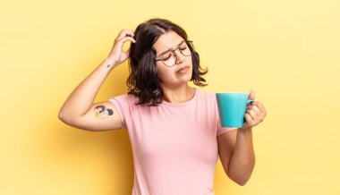 Genç İspanyol kadın şaşkın ve kafası karışık, kaşınan bir kafa hissediyor. kahve fincanı konsepti
