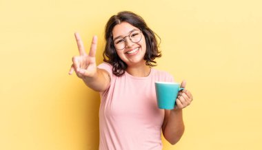 Genç İspanyol kadın gülümsüyor ve mutlu görünüyor, zafer ya da barış işareti yapıyor. kahve fincanı konsepti