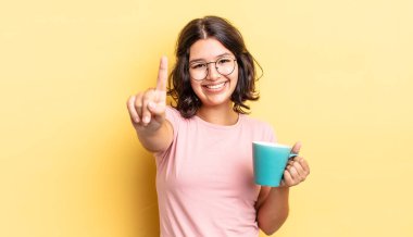 Genç İspanyol kadın gülümsüyor ve arkadaş canlısı görünüyor, bir numarayı gösteriyor. kahve fincanı konsepti