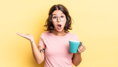 Genç İspanyol kadın şaşırmış ve şok olmuş görünüyor. Çenesi düşmüş ve elinde bir nesne tutuyor. kahve fincanı konsepti
