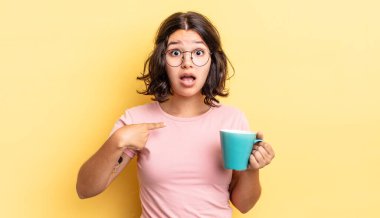 Genç İspanyol kadın şok olmuş ve ağzı açık, kendini işaret ederek şaşırmış görünüyor. kahve fincanı konsepti