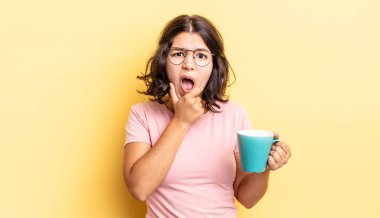 Ağzı ve gözleri sonuna kadar açık ve eli çenesinde olan genç İspanyol kadın. kahve fincanı konsepti