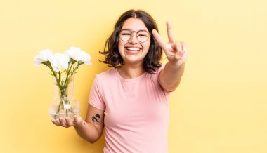 Genç İspanyol kadın gülümsüyor ve mutlu görünüyor, zafer ya da barış işareti yapıyor. çiçek saksı konsepti