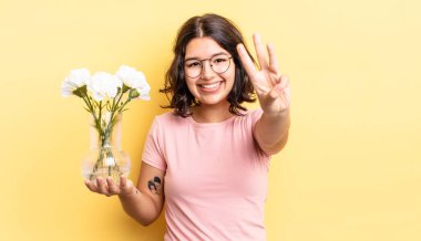 Genç İspanyol kadın gülümsüyor ve arkadaş canlısı görünüyor, üç numarayı gösteriyor. çiçek saksı konsepti