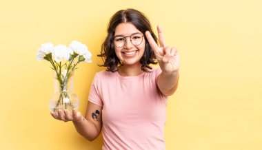 Genç İspanyol kadın gülümsüyor ve arkadaş canlısı görünüyor, iki numarayı gösteriyor. çiçek saksı konsepti