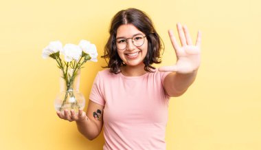 Genç İspanyol kadın gülümsüyor ve arkadaş canlısı görünüyor, beş numarayı gösteriyor. çiçek saksı konsepti