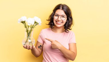 Genç İspanyol kadın neşeyle gülümsüyor, mutlu hissediyor ve bir tarafı işaret ediyor. çiçek saksı konsepti