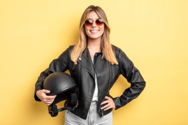 Güzel sarışın kız mutlu bir şekilde gülümsüyor, eli kalçasında ve kendinden emin. Motosiklet kaskı konsepti