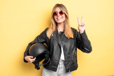 Güzel sarışın kız gülümsüyor ve arkadaş canlısı görünüyor. İki numarayı gösteriyor. Motosiklet kaskı konsepti