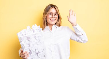 Güzel sarışın kız mutlu bir şekilde gülümsüyor, el sallıyor, hoş geldin diyor ve seni selamlıyor. kağıt toplar çöp konsepti