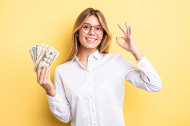 Güzel sarışın kız kendini mutlu hissediyor, iyi bir jestle onaylıyor. Dolar banknotları konsepti