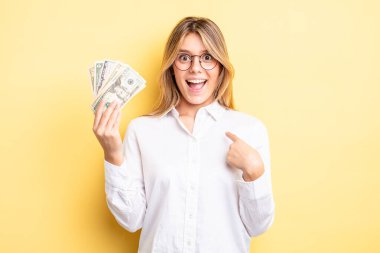 Güzel sarışın kız mutlu hissediyor ve heyecanla kendini işaret ediyor. Dolar banknotları konsepti