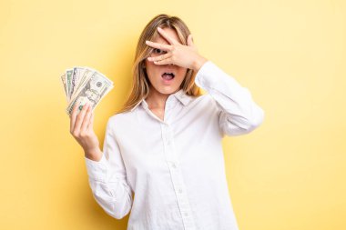 Güzel sarışın kız şok olmuş, korkmuş ya da korkmuş görünüyor. Yüzü elle kapatılmış. Dolar banknotları konsepti