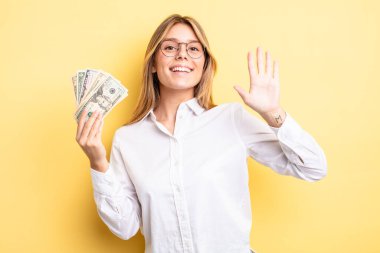 Güzel sarışın kız mutlu bir şekilde gülümsüyor, el sallıyor, hoş geldin diyor ve seni selamlıyor. Dolar banknotları konsepti