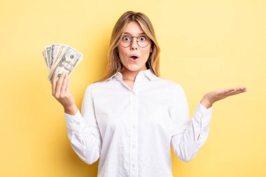 Güzel sarışın kız şaşırmış ve şok olmuş görünüyor. Çenesi düşmüş ve elinde bir nesne var. Dolar banknotları konsepti